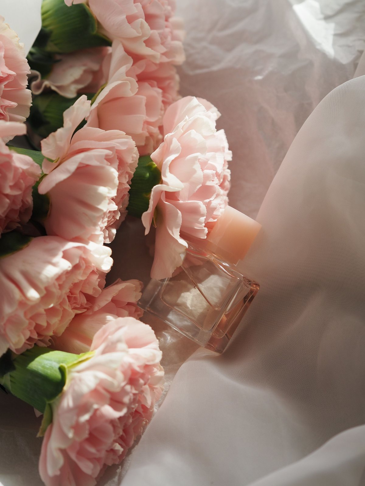 Clear perfume bottle styled beside pale pink flowers and white fabric.