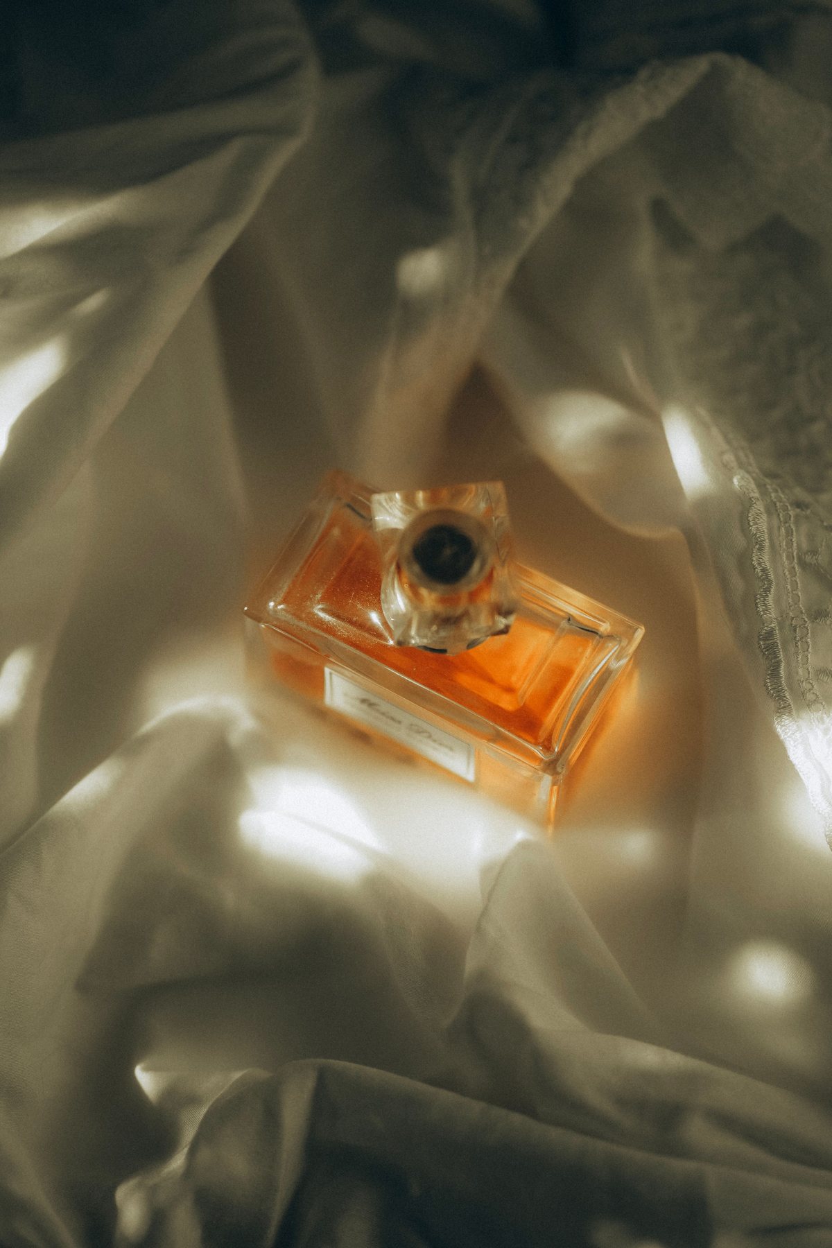 Amber perfume bottle glowing softly on cream fabric in filtered light.