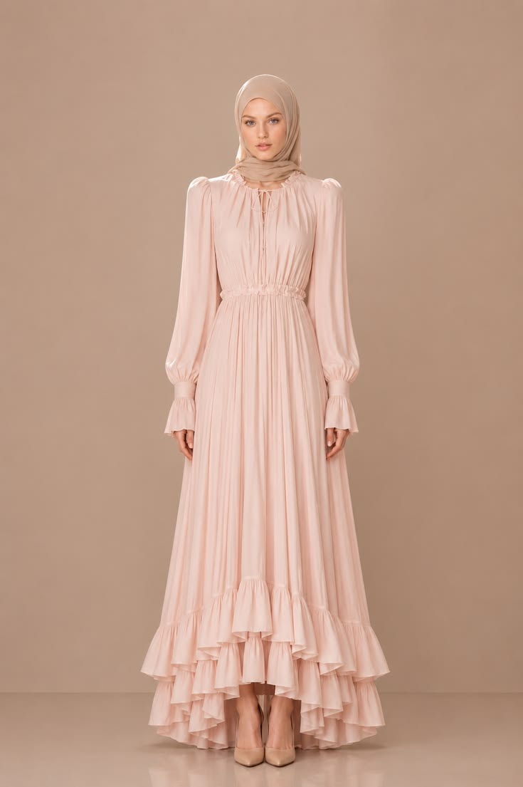 Blush pink modest wedding guest dress with long sleeves, gathered waist, high-low layered ruffle hem, taupe hijab, and nude pumps.